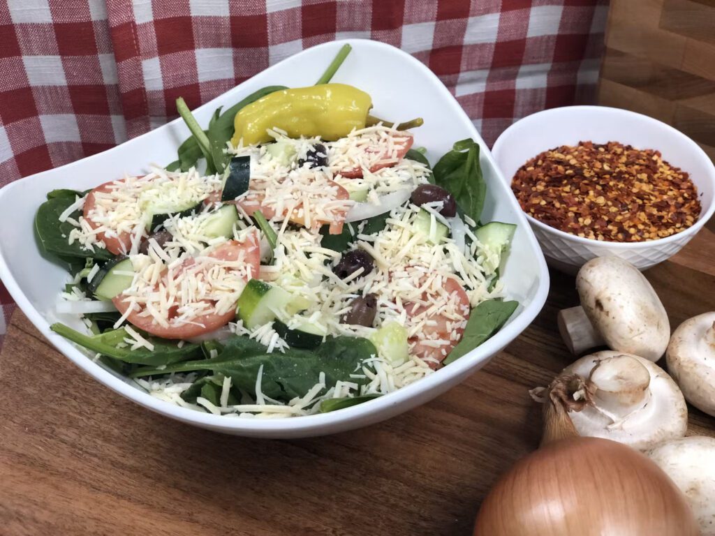 Greek salad at Manny & Olga's Salad bowl with fresh veggies, cheese, paneer and meat, placed on a wooden board with chilli flakes, mushrooms and an onion placed next to it.