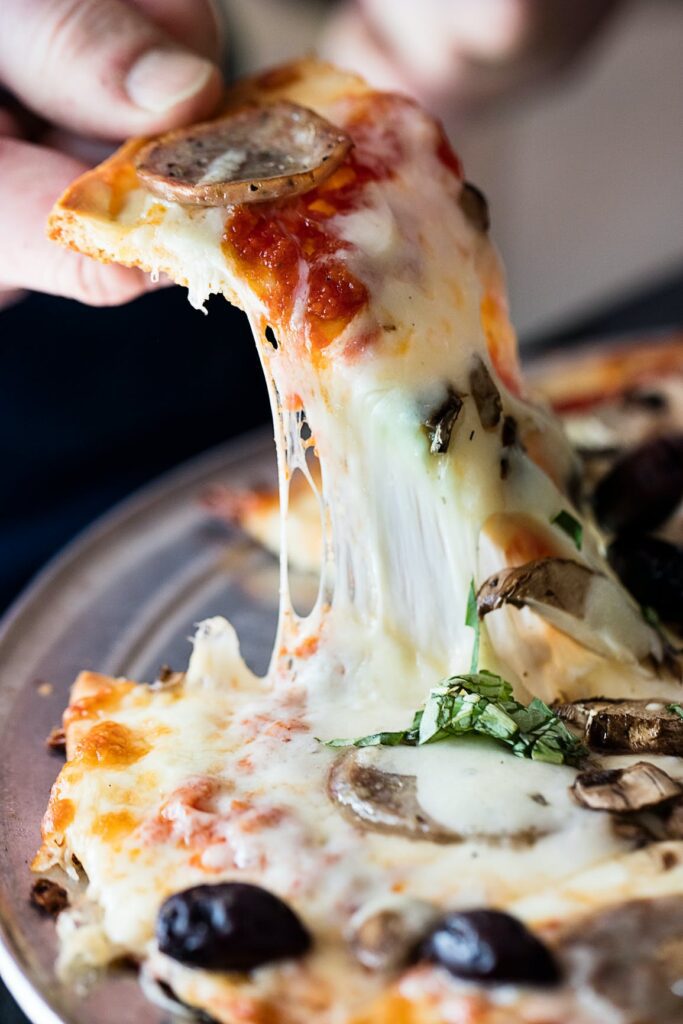 Hand lifting a cheesy slice of mushroom and olive pizza with stringy mozzarella and sliced sausage.