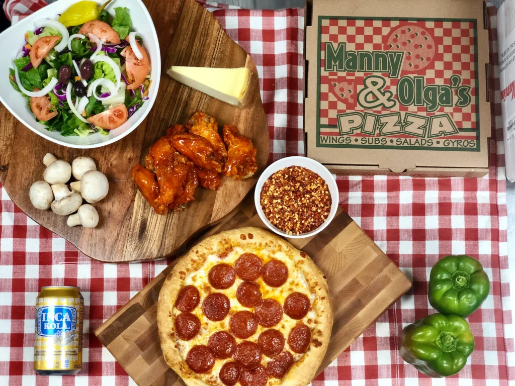 Assorted Manny & Olga’s Pizza items including pepperoni pizza, buffalo wings, fresh salad, mushrooms, and green peppers on a red checkered tablecloth.