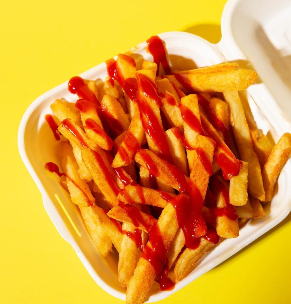 A white takeout container filled with golden French fries drizzled with red ketchup, placed on a yellow background.
