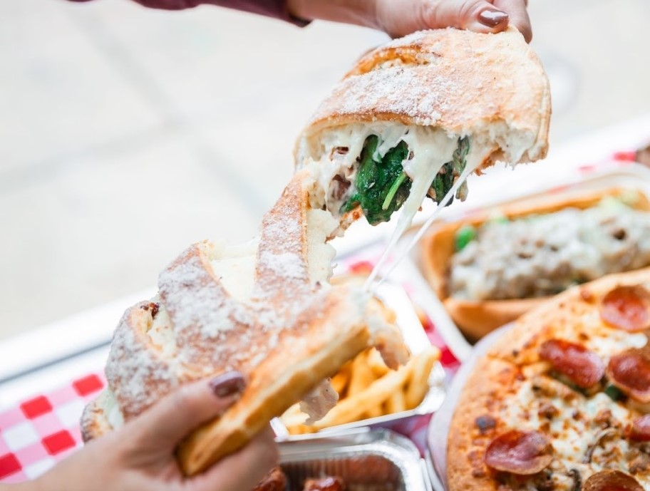 Hands pulling apart a cheesy spinach-filled calzone with sides of pizza, wings, fries, and toasted ravioli on a checkered table.