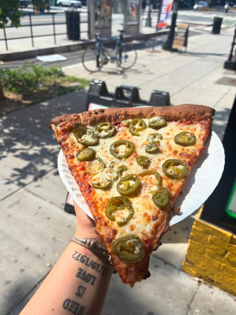 Fresh jalapeño pizza slice from Manny & Olga’s held outdoors on a sunny Georgetown sidewalk.