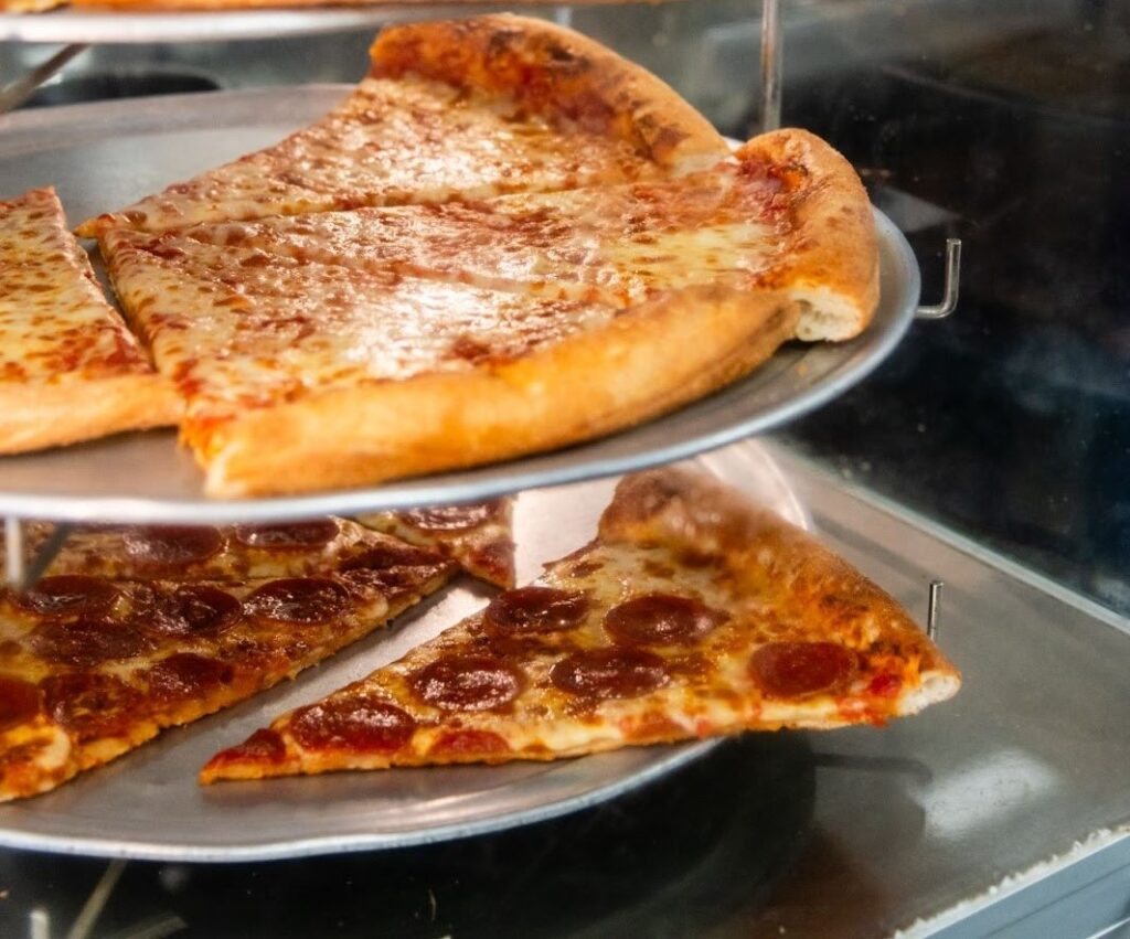 Display case with trays of cheese and pepperoni pizza slices stacked in a heated rack.