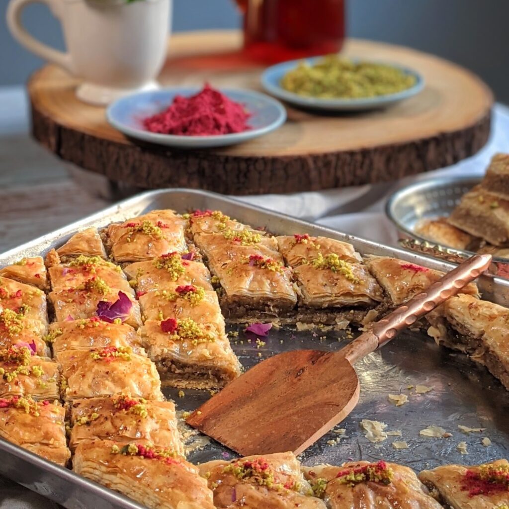 A tray of freshly baked baklava A tray of freshly baked baklava cut into diamond shapes, topped with crushed pistachios and dried rose petals, with a copper serving spatula placed on the tray.