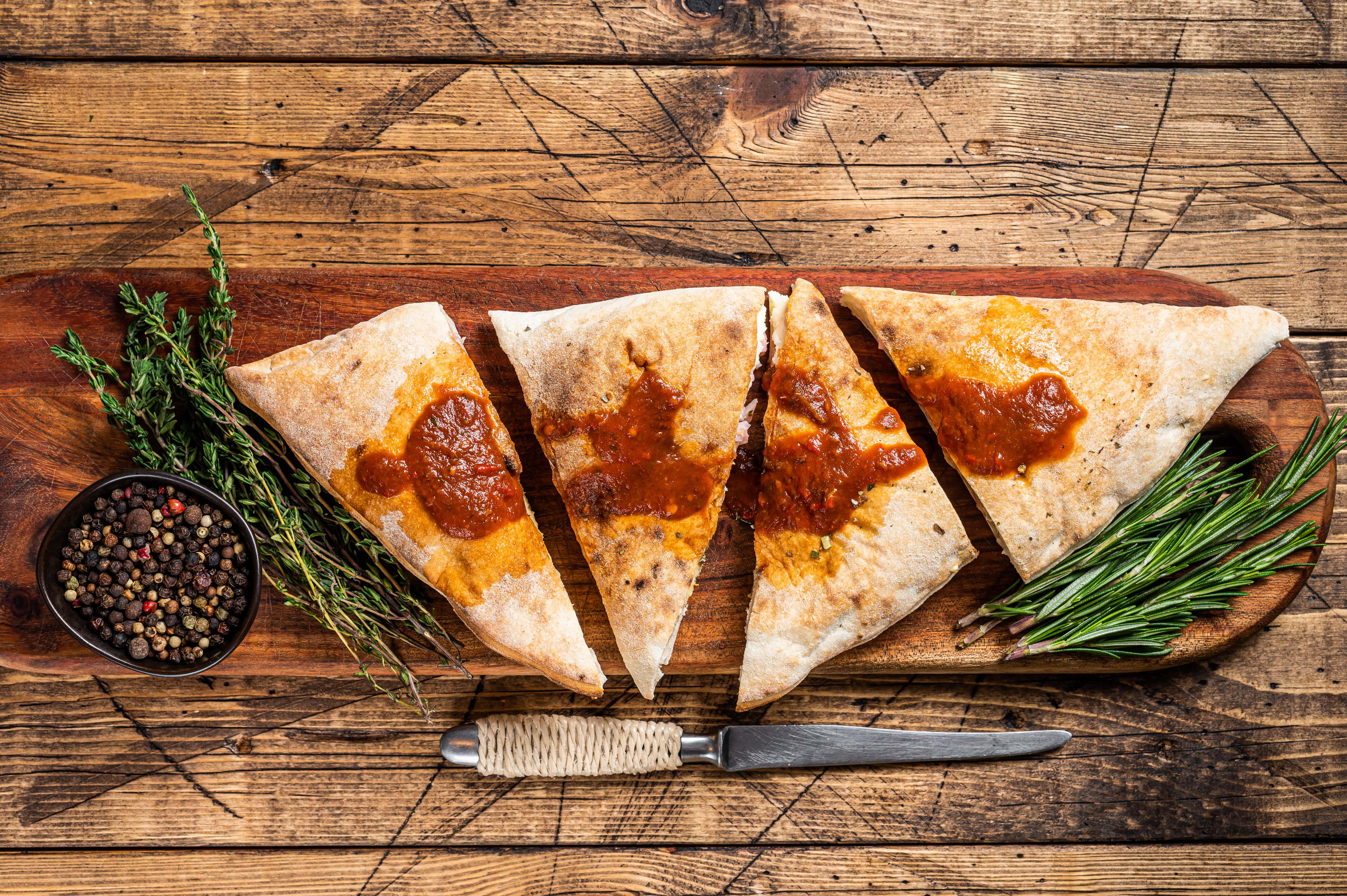 Calzone and pizza slices on a wooden board with herbs and spices.