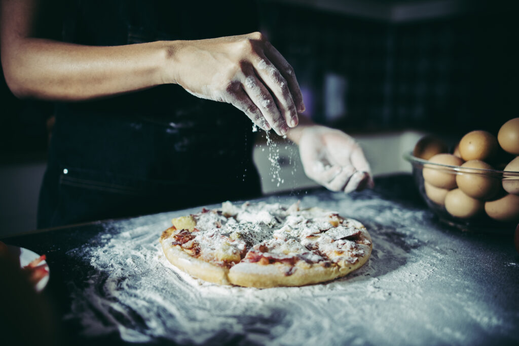 Chef hands pouring flour powder on raw dough, like at Manny & Olga’s kitchen.