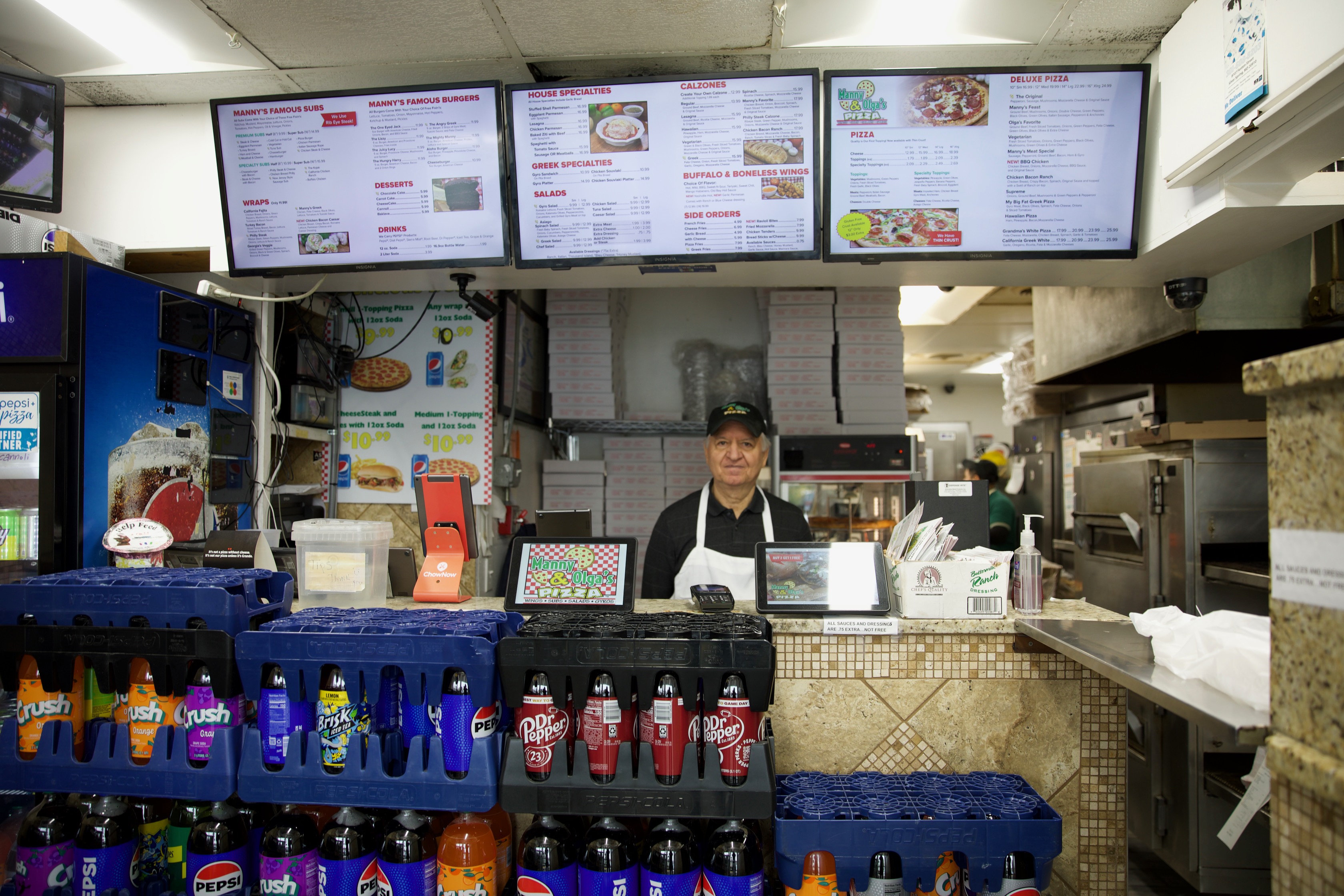 Manny & Olga’s counter with menu boards, staff preparing orders, and drinks displayed for customers.