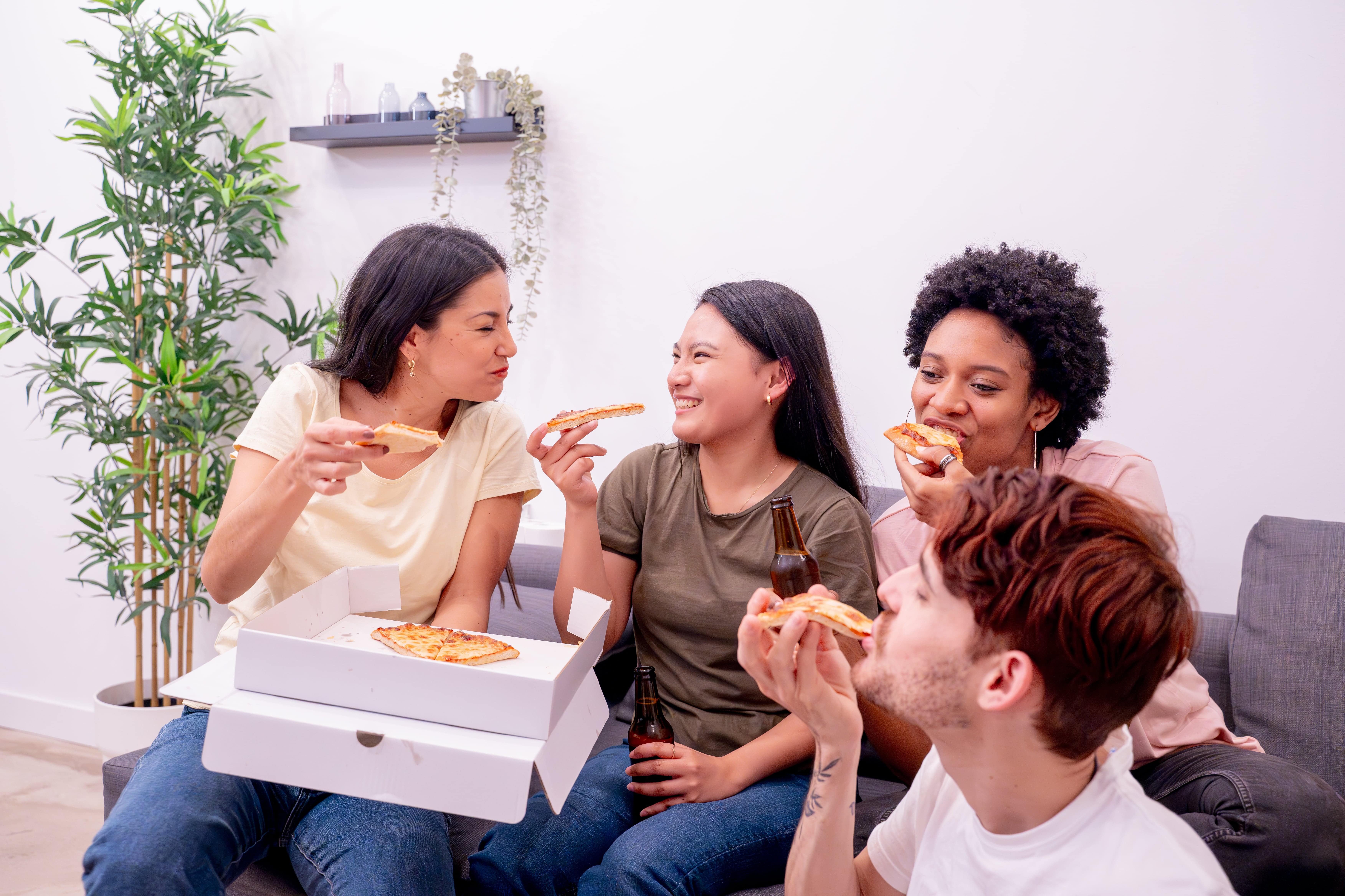 A group of four friends enjoying a pizza party A group of four friends enjoying a pizza party in a cozy living room, smiling and chatting while eating slices of pizza with drinks.