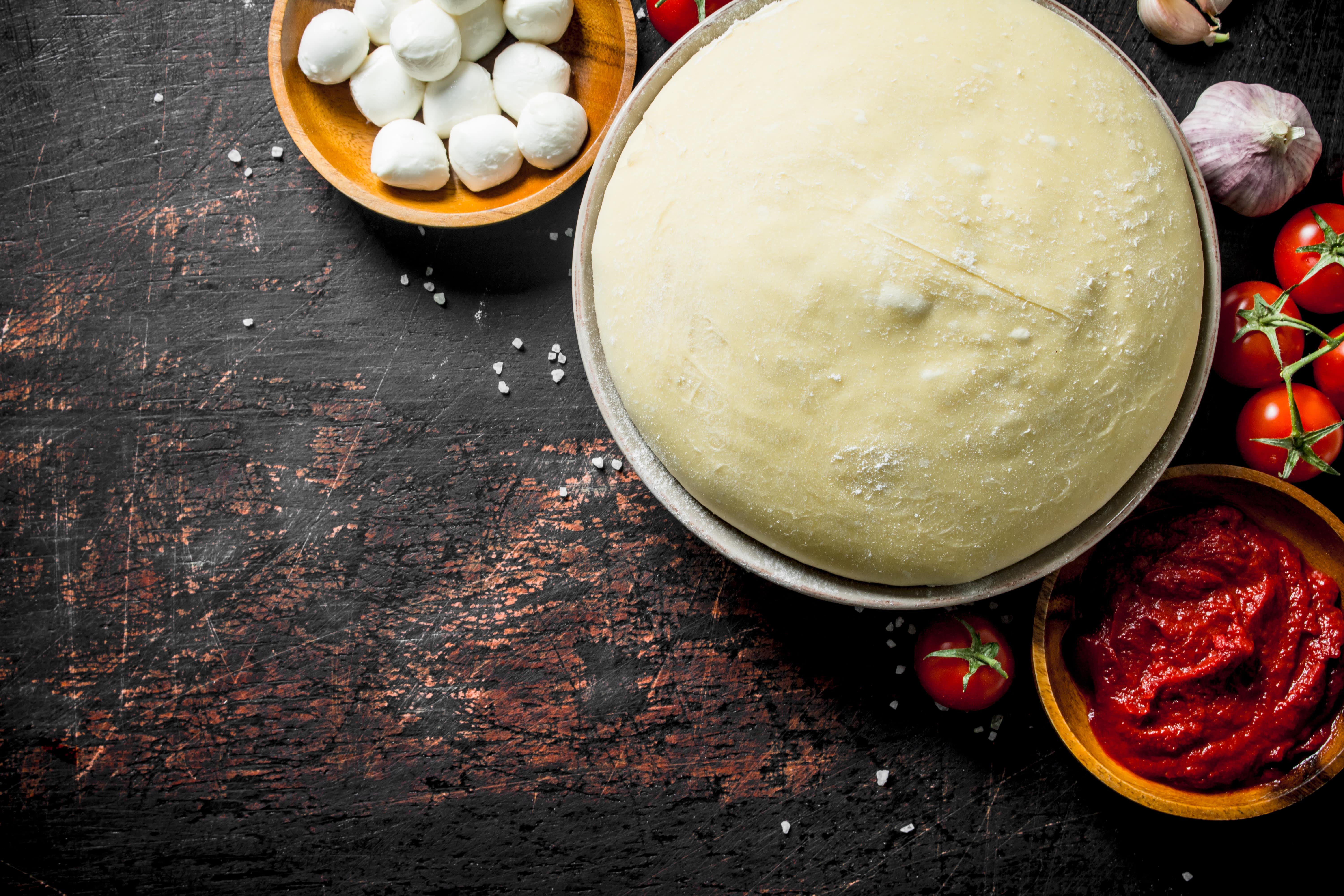 Raw pizza dough with mozzarella, tomato paste, cherry tomatoes, garlic, and coarse salt on a rustic dark surface.