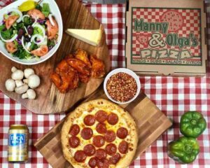 A catering spread from Manny & Olga's Pizza with a pepperoni pizza, chicken wings, salad, mushrooms, peppers, a can of soda, and red pepper flakes.