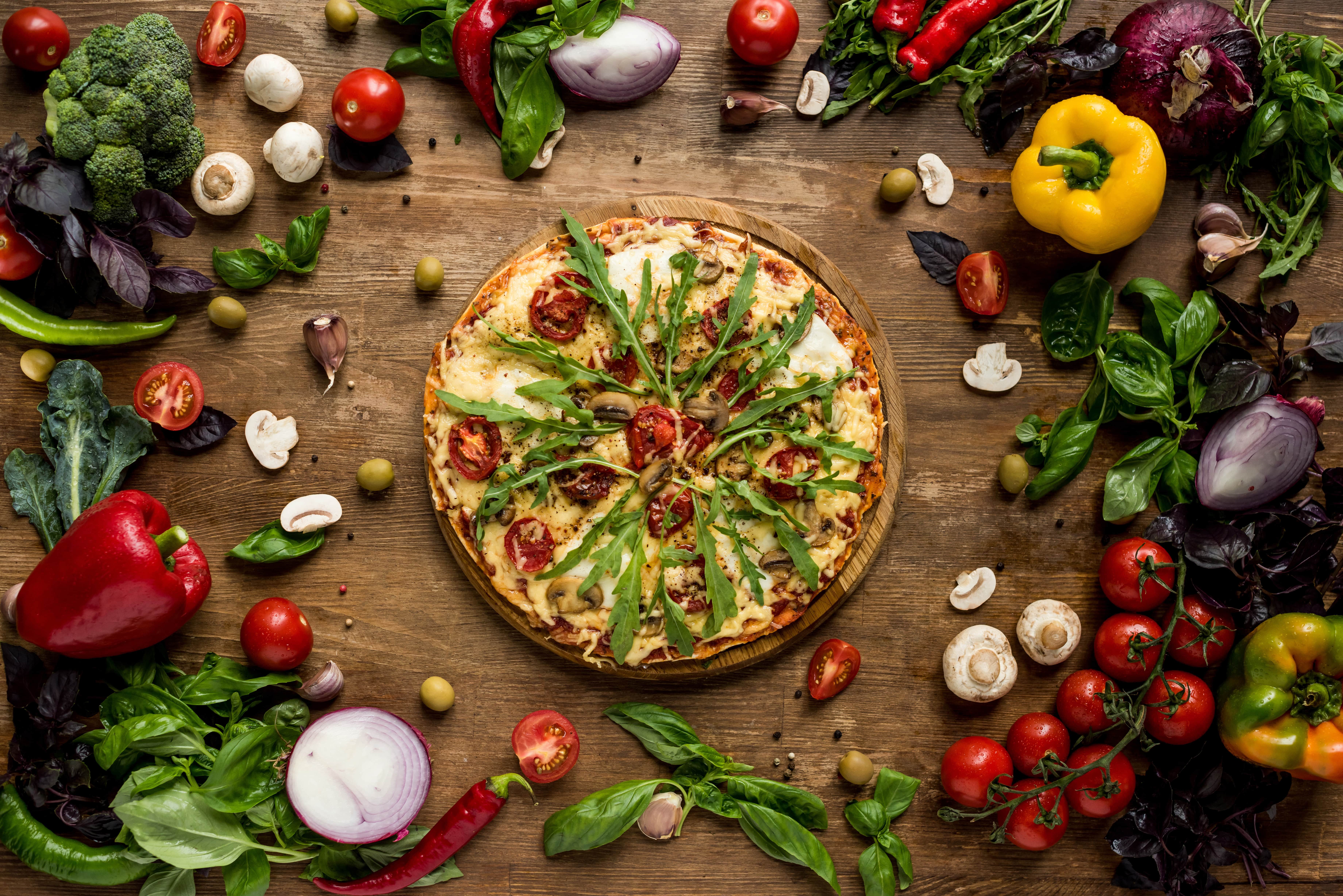 A top-down view of a pizza with various toppings A top-down view of a pizza with various toppings, surrounded by fresh vegetables like tomatoes, mushrooms, onions, and peppers. The pizza and vegetables are arranged on a wooden table
