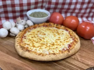 Whole Manny & Olga’s cheese pizza on a wooden board with fresh tomatoes, mushrooms, and sauce on a checkered tablecloth.