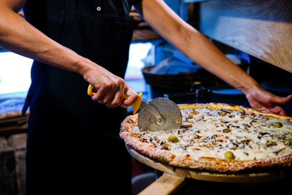 Chef slicing a large cheesy pizza fresh out of the oven using a pizza cutter in a rustic kitchen setting.
