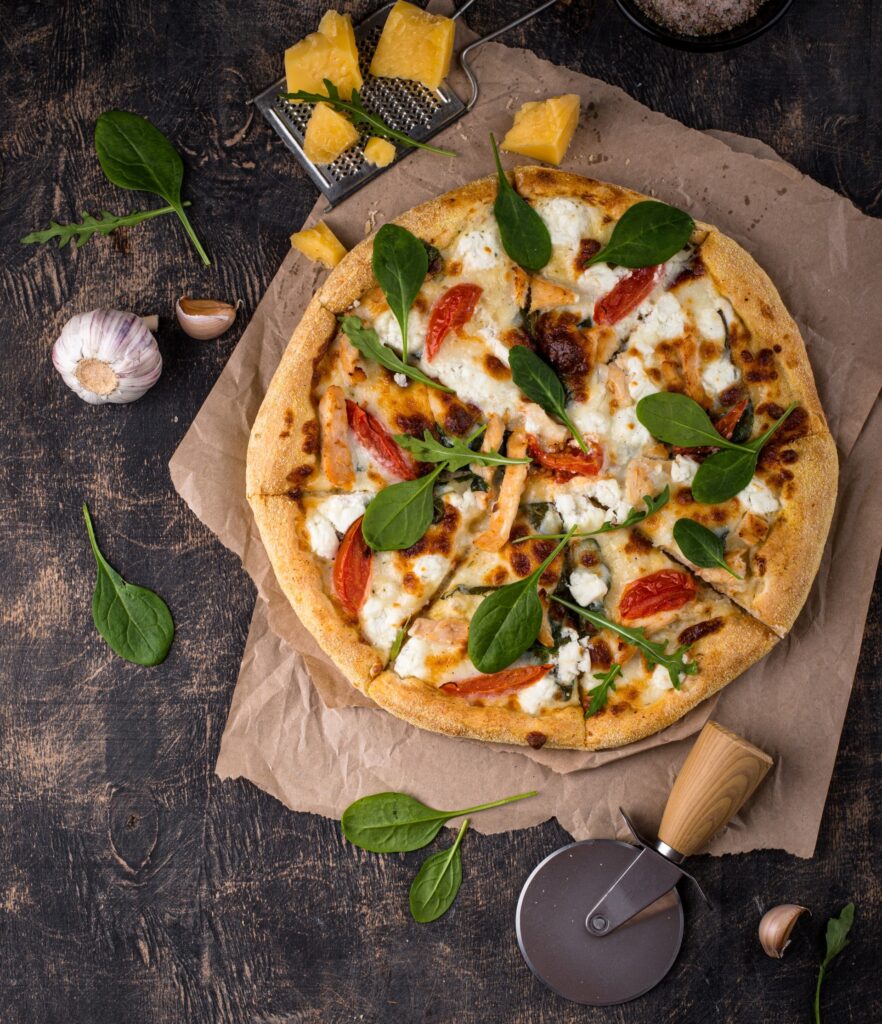 Top view of a fresh veggie pizza on rustic paper, surrounded by garlic, cheese, and a pizza cutter.