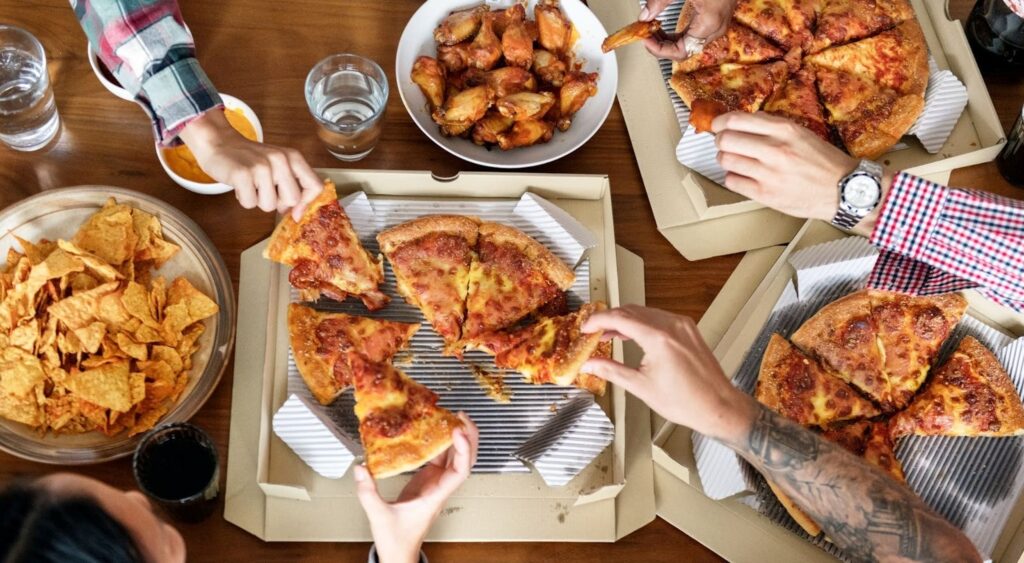 A group of friends gathered around a table enjoying pizza A group of friends gathered around a table, enjoying a lively pizza party. The table is spread with delicious food, including pizza slices, nachos, chicken wings, drinks, and dipping sauces.