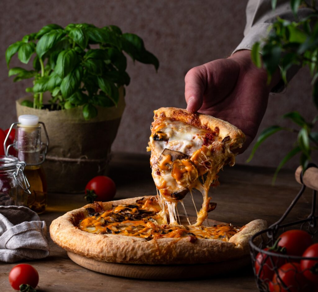 Hand lifting a cheesy slice of mushroom pizza from a wooden board, surrounded by fresh basil, tomatoes, and olive oil.