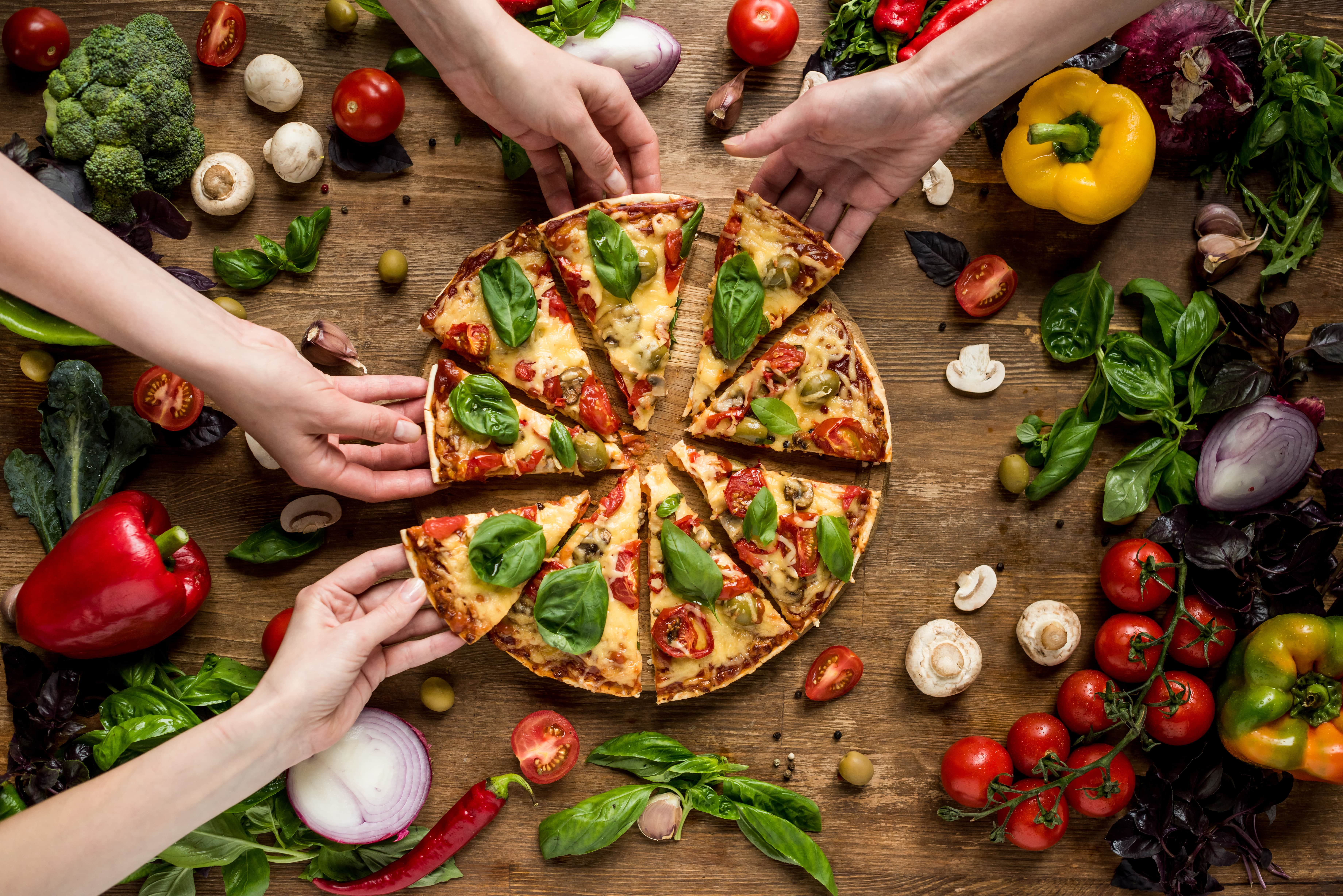 A delicious pizza with fresh basil A delicious pizza with fresh basil, surrounded by a variety of colorful vegetables and hands reaching for slices.