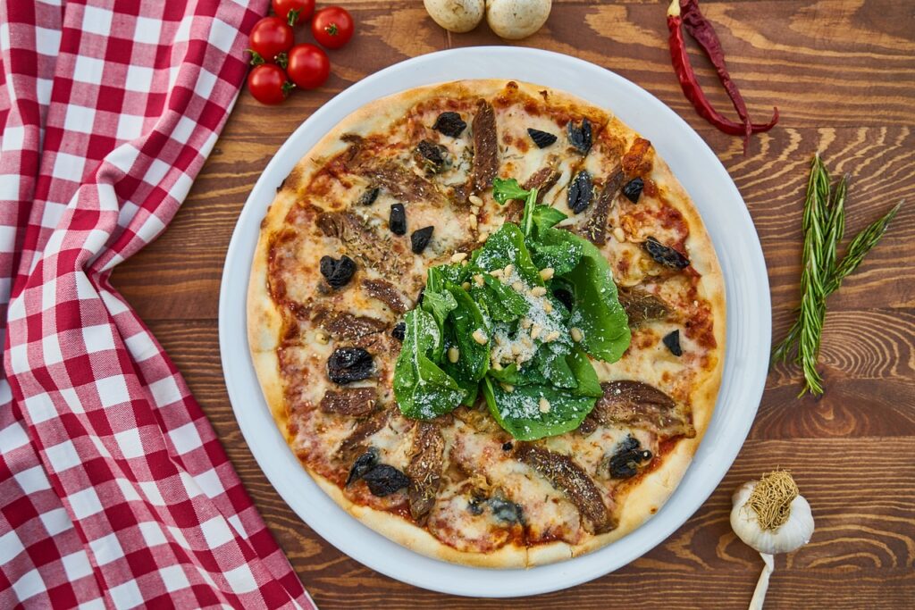A freshly baked pizza topped with caramelized onions, black olives, and a generous handful of fresh spinach leaves sprinkled with grated Parmesan cheese, served on a white plate. The wooden table is adorned with a red-and-white checkered cloth, cherry tomatoes, garlic, rosemary, and dried chili, adding a rustic and appetizing touch to the scene for a movie night.