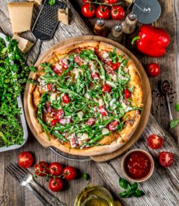 Fresh veggie pizza with cherry tomatoes, bell peppers, and greens on a wooden table.