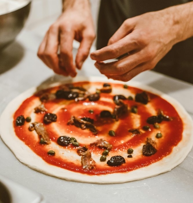 Skilled chef crafting a delicious pizza with fresh, flavorful toppings! Hands adding toppings to fresh pizza dough with tomato sauce, olives, capers, and anchovies for a romantic homemade dinner.