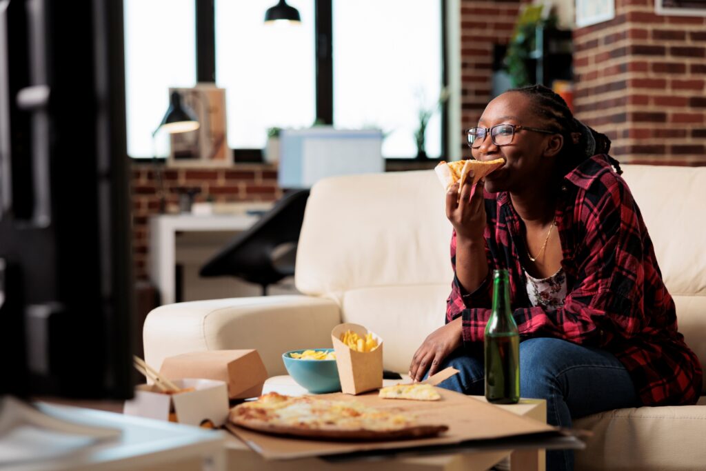 A woman wearing a red striped t-shirt, watching TV, eating pizza at night.