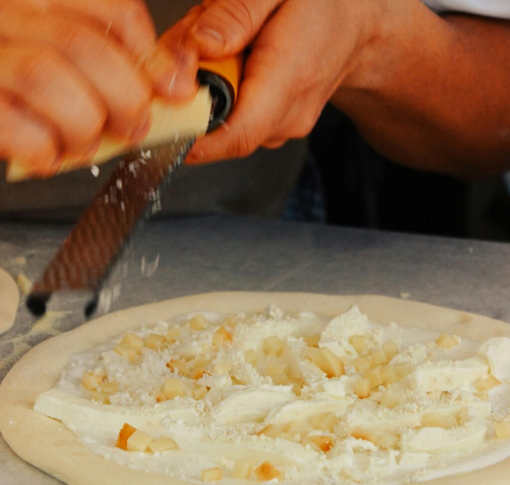 Fresh cheese grated over pizza sauce and toppings at Manny & Olga’s using locally sourced dairy.