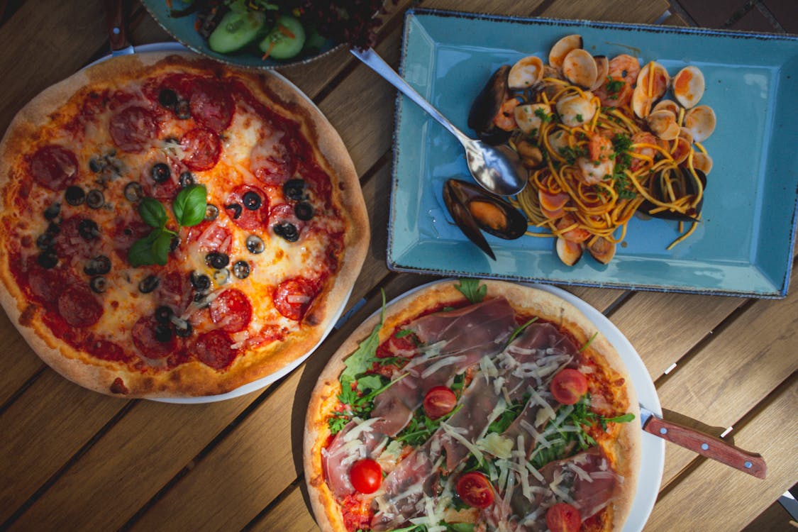 Overhead shot of two artisan pizzas with olives, arugula, prosciutto, and seafood pasta, celebrating local Italian cuisine.