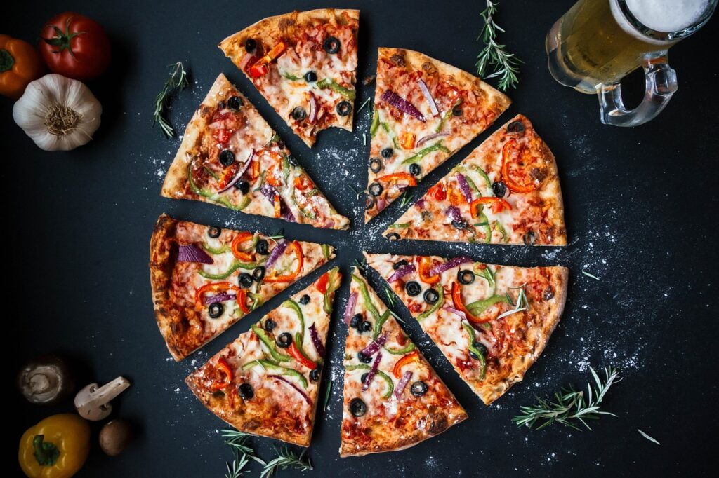 Sliced pizza with red onions, bell peppers, and olives surrounded by colorful, fresh vegetables at Manny & Olga’s.