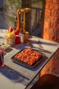 A square pizza topped with pepperoni slices served on a black plate with fries and condiments on the table.