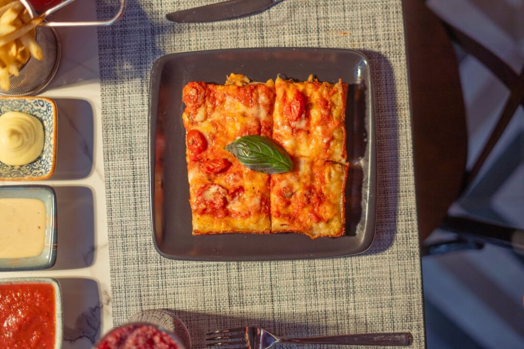 A square pizza topped with tomatoes and a basil leaf on a black plate, with sauces and fries on the side.