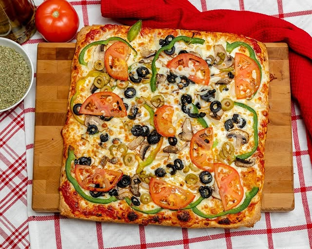 A square pizza with various toppings, including mushrooms, onions, green peppers, olives, and more, on a wooden board.