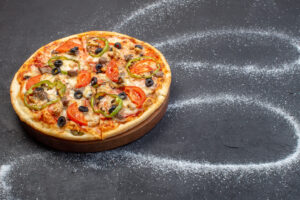 Pizza on a wooden board with olives and peppers, surrounded by flour swirls resembling a galaxy pattern.