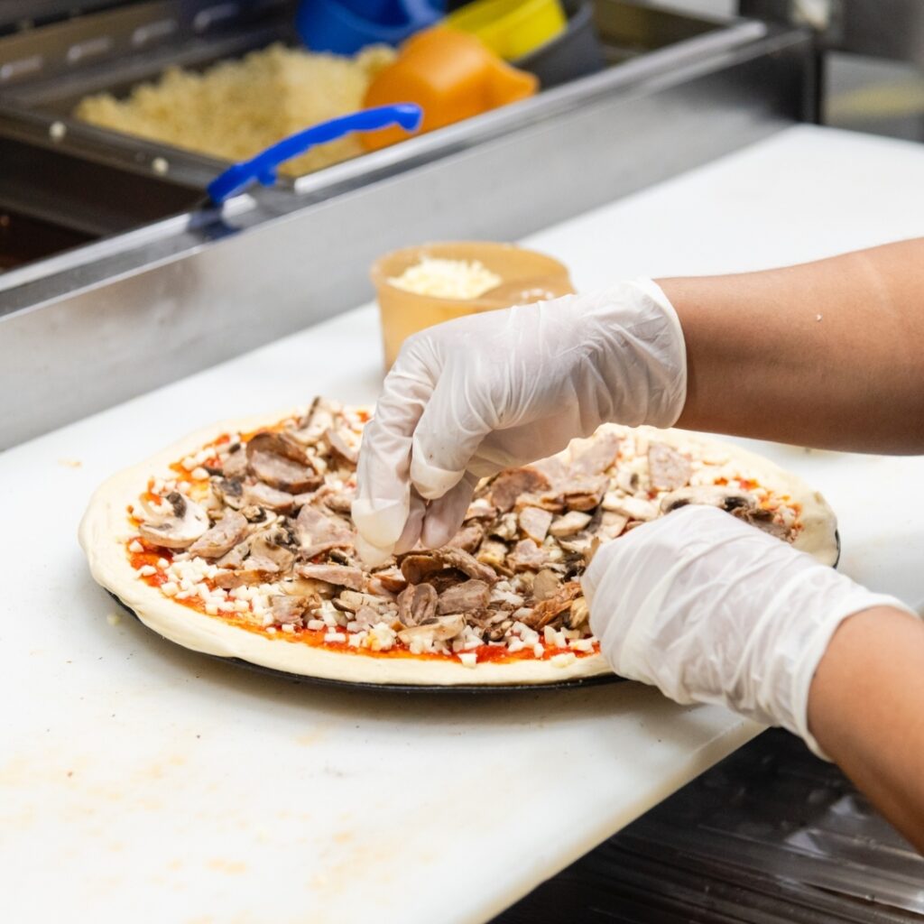 Manny & Olga’s staff member adding meat and mushrooms to a pizza in the kitchen prep area.