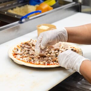 Manny & Olga’s staff member adding meat and mushrooms to a pizza in the kitchen prep area.