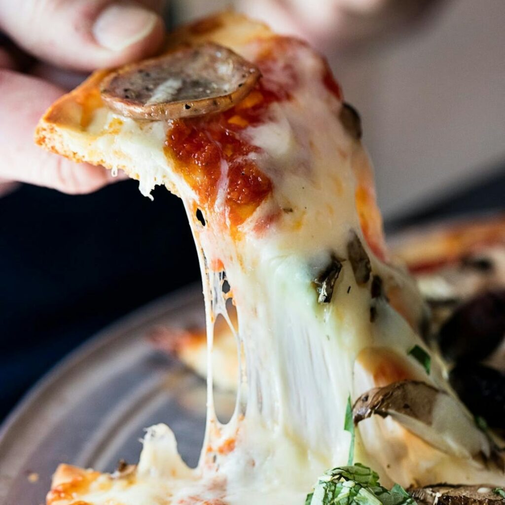 Manny & Olga’s mushroom pizza slice lifted with a gooey cheese pull, capturing a viral food moment perfect for social media.