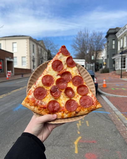 A hand holding a slice of pepperoni pizza with a crisp crust, set against a street view with brick buildings and clear skies.