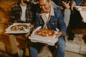 Friends sitting on steps enjoying hot cheesy pizzas in boxes at night.