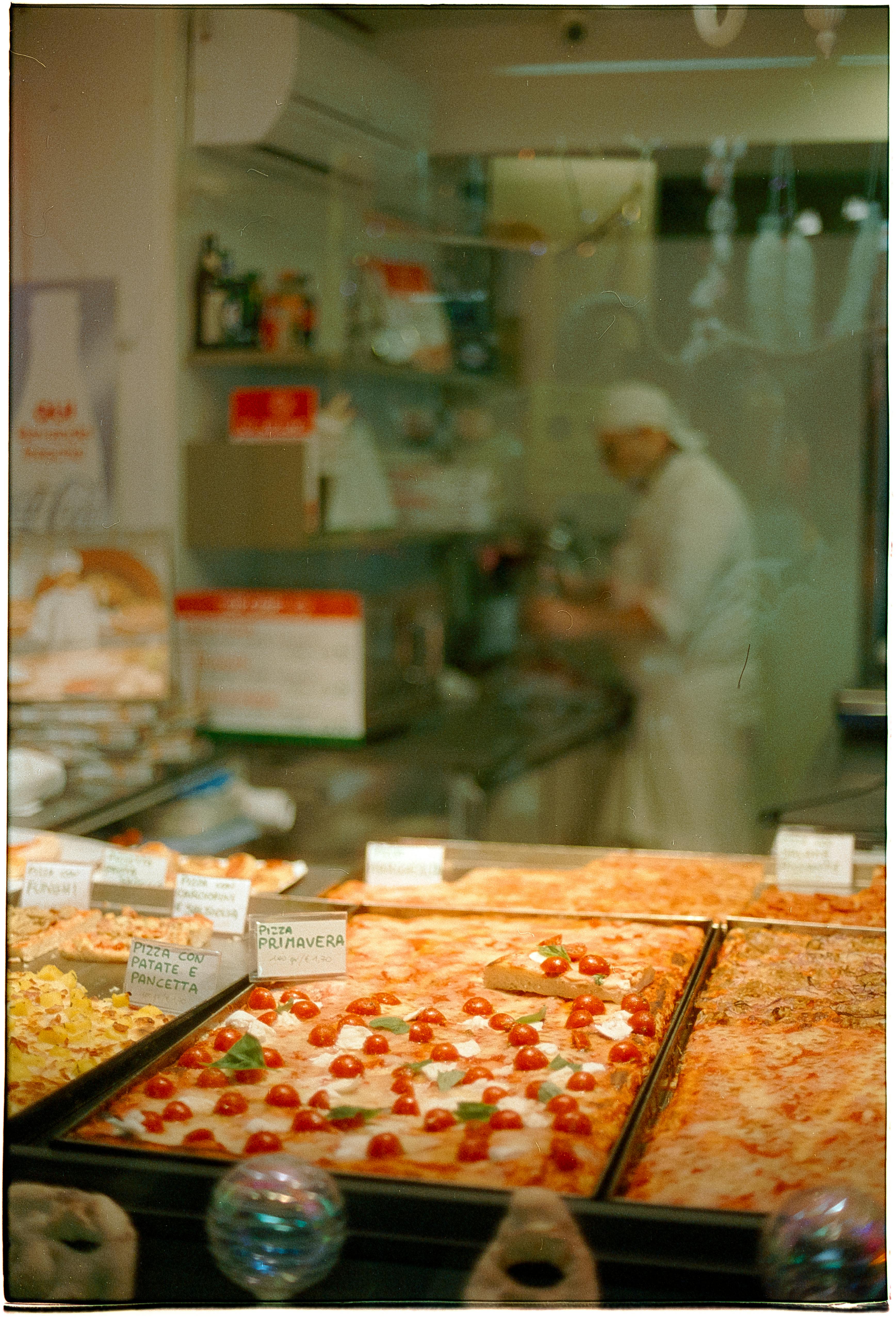 Pizzeria window display with assorted pizzas and chef preparing dough in the background.