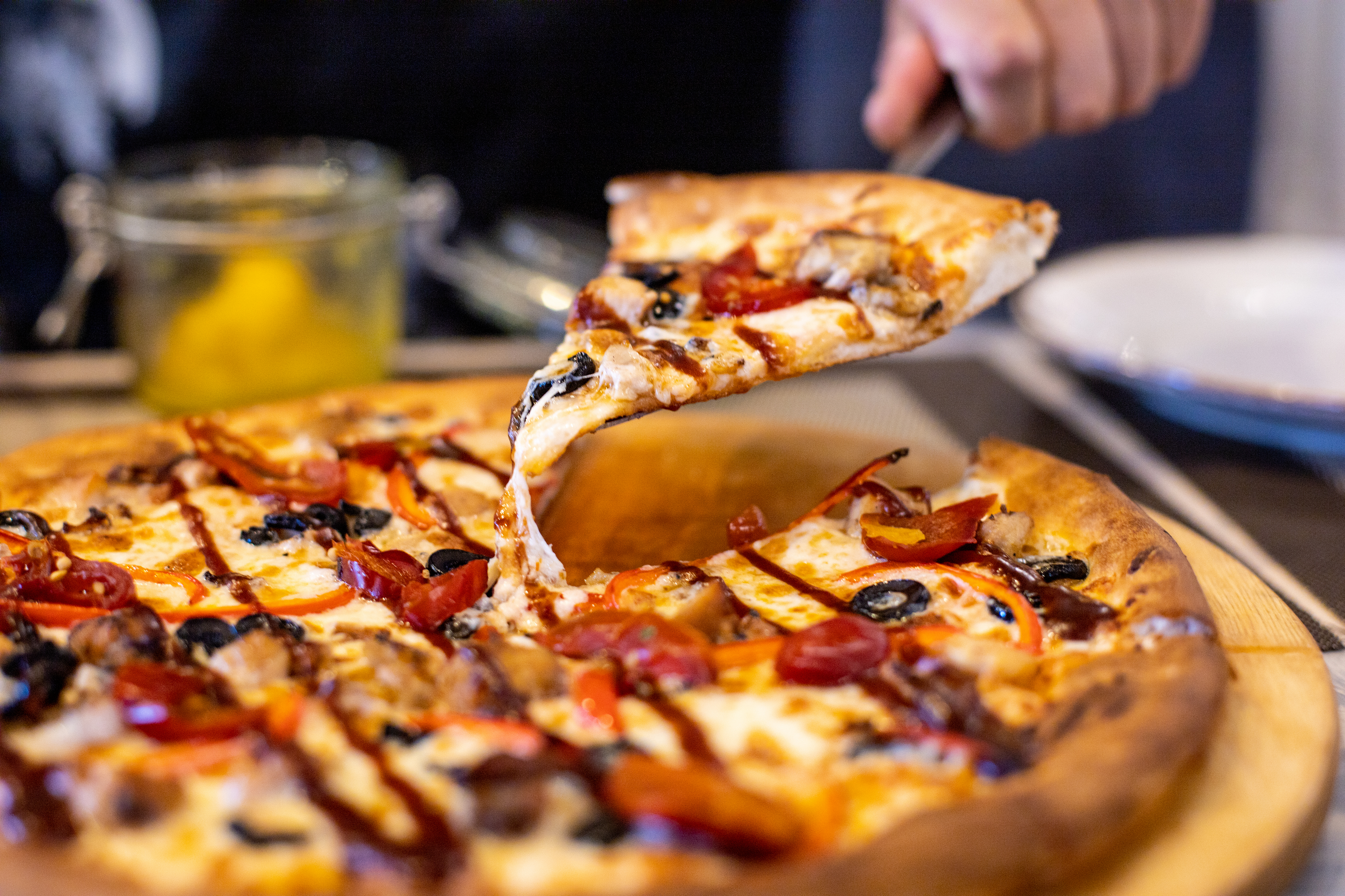 Hand lifting a cheesy pizza slice topped with olives, peppers, and sausage from a wooden tray.