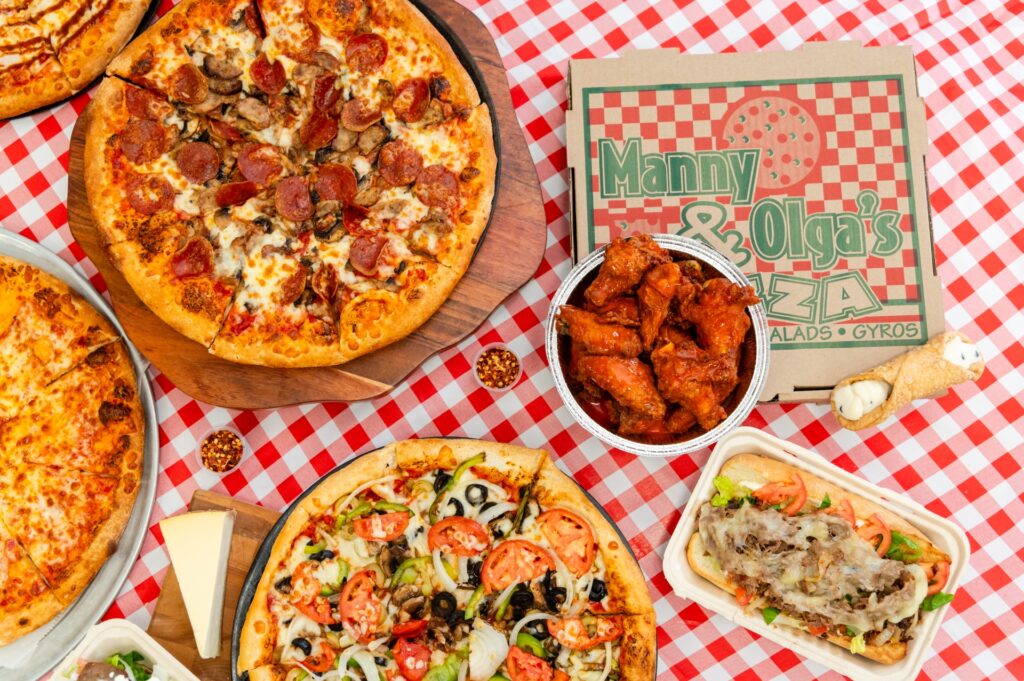 Assorted pizzas, wings, and subs spread on a table showcasing Manny & Olga’s catering favorites.
