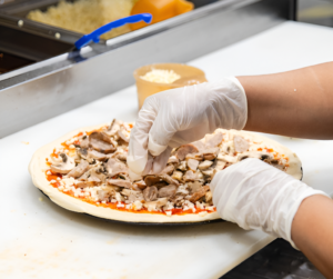 Fresh pizza being topped with mushrooms and meat by hand at Manny & Olga’s kitchen.