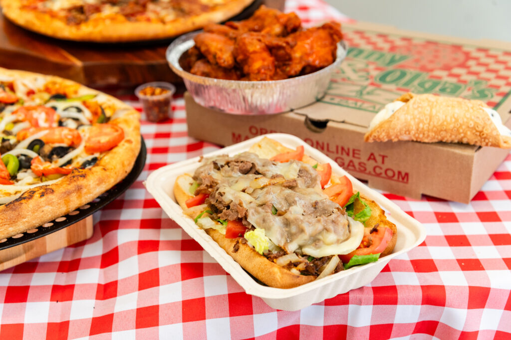 Loaded steak and cheese sub with pizzas and wings spread on the table from Manny & Olga’s.