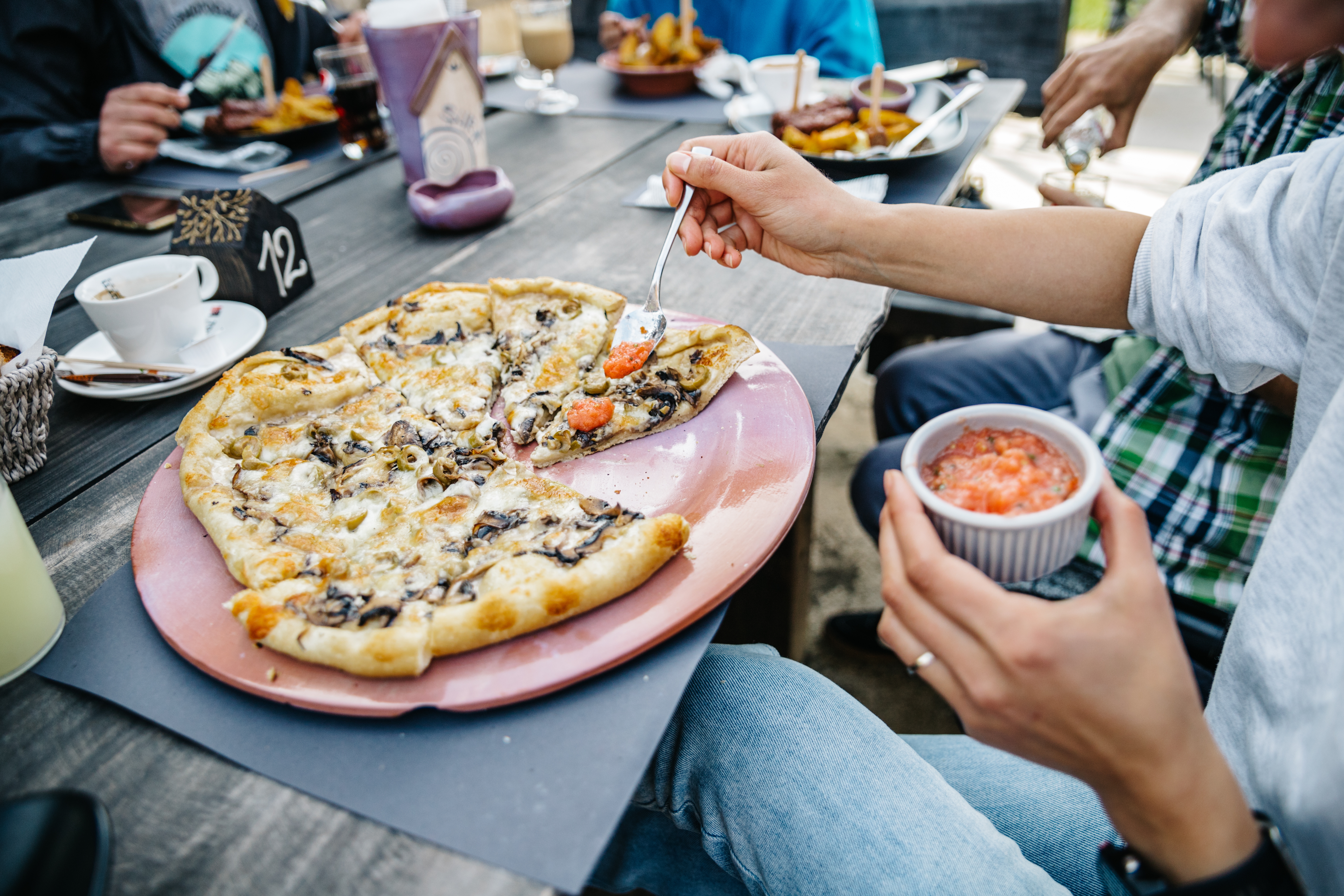 Person adding salsa to a mushroom and olive pizza slice at an outdoor restaurant table with friends.