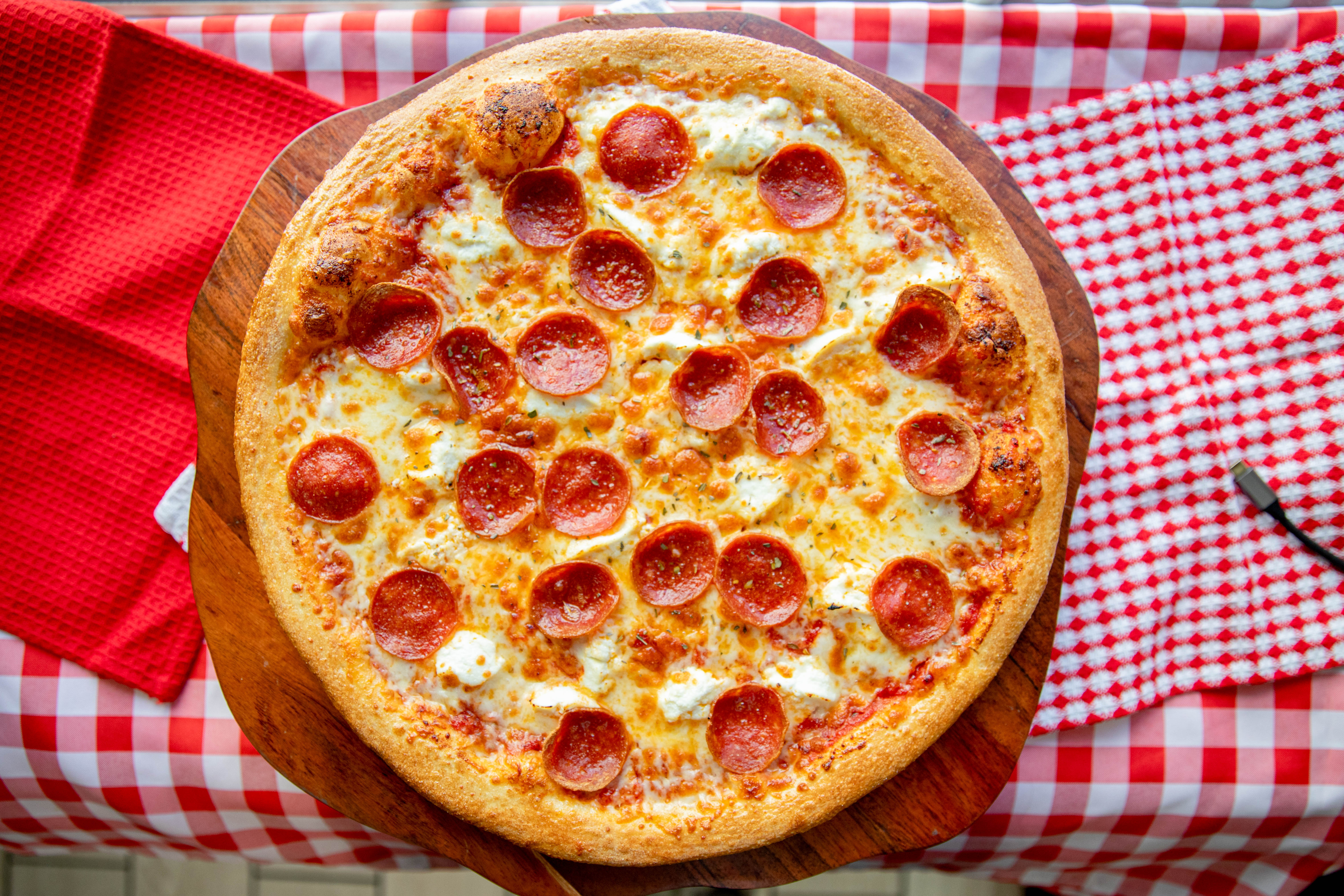 Manny & Olga’s pepperoni pizza with melted mozzarella on a wooden board over a red checkered tablecloth.