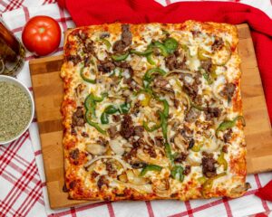 Manny and Olga’s square steak and veggie pizza topped with peppers, onions, and melted cheese on a wooden board.