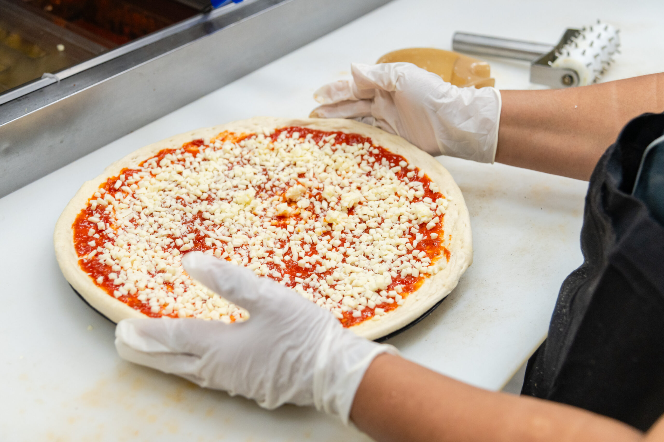 Gloved hands preparing a Manny & Olga’s cheese pizza with sauce and shredded cheese.