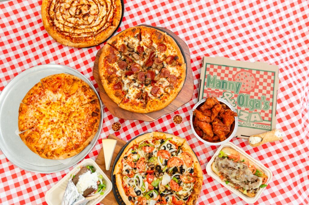 Manny and Olga’s pizza spread featuring pepperoni and veggie pizzas, wings, and a loaded sub arranged on a red checkered tablecloth.