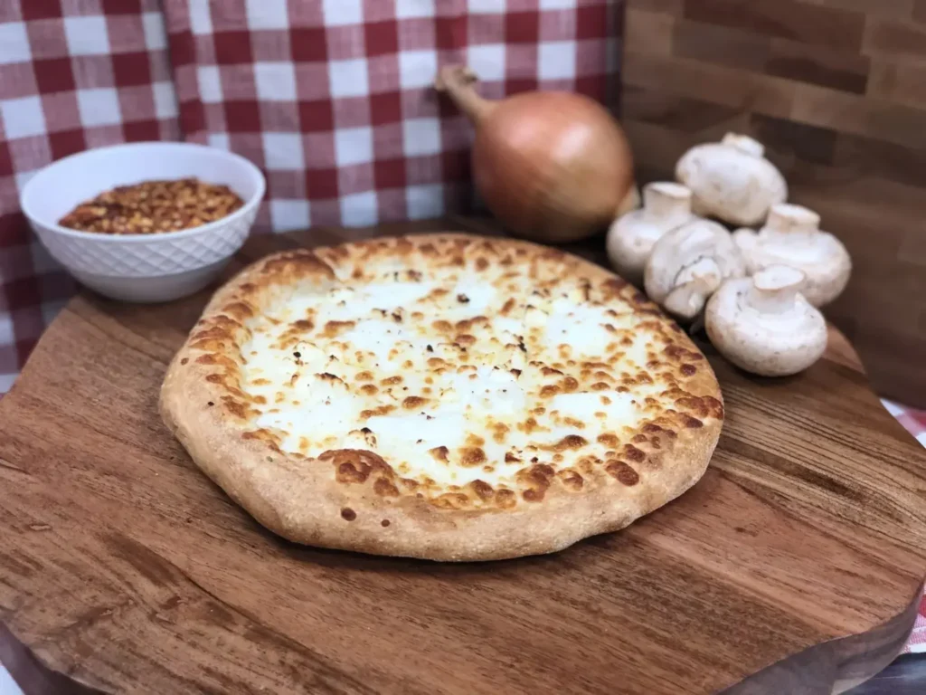 Freshly baked pizza topped with vegetables and spices served on a wooden board with red pepper flakes and onions on the side.