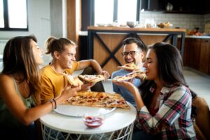 Four friends enjoying pizza indoors, laughing around a table.