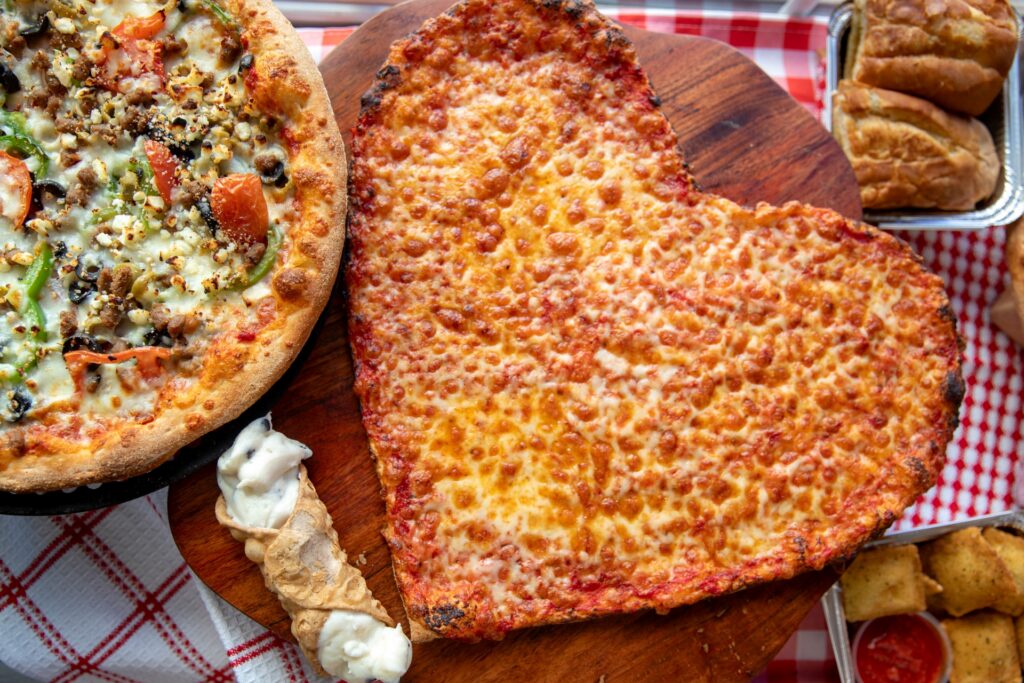 A heart-shaped cheese pizza from Manny & Olga’s Pizza served on a wooden board with specialty pies and sides.